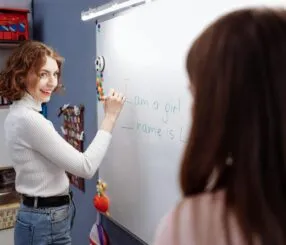 Mulher escreve em um quadro branco enquanto sorri, ensinando uma criança.